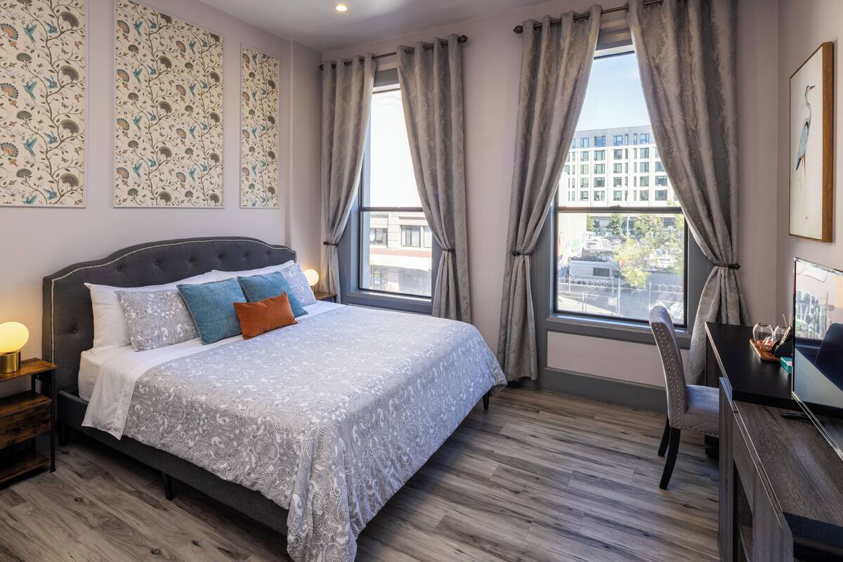 A hotel room with a bed, bedside table, lamp, chair, desk, and large windows with curtains.