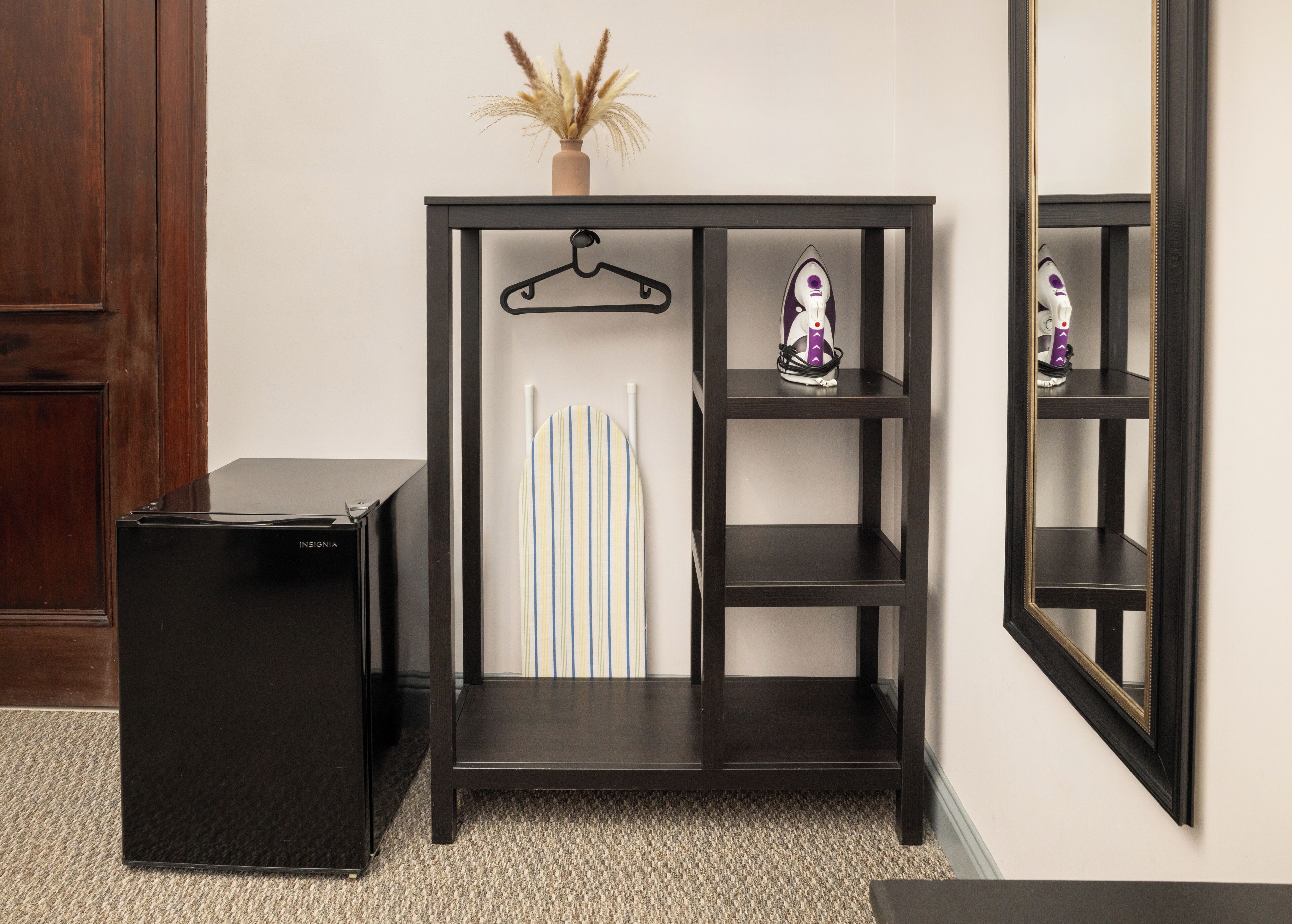 A black console table with a hanging garment, a small shelf with an iron, and a vase with dried plants.