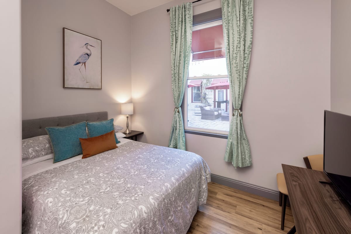 A bedroom with a bed, a desk, a TV, a lamp, and a window with curtains.