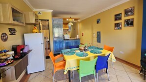 A compact kitchen with a refrigerator, microwave, and coffee machine. A dining table with colorful chairs is in the center. - Country View Apartment (Mellieha)