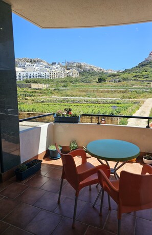 A balcony with a table and chairs, potted plants, and a view of a hillside with buildings and greenery. - Country View Apartment (Mellieha)