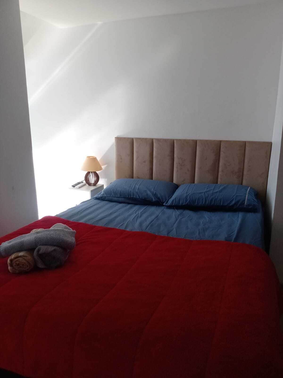 Una cama bien hecha con colcha roja y azul, un cabecero marrón y una lámpara en una mesita de noche.