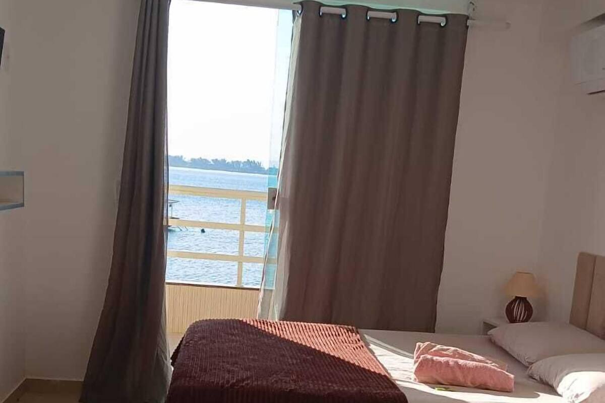 Ένα υπνοδωμάτιο με ένα κρεβάτι, ένα κομοδίνο, ένα φωτιστικό και θέα στη θάλασσα από ένα παράθυρο.