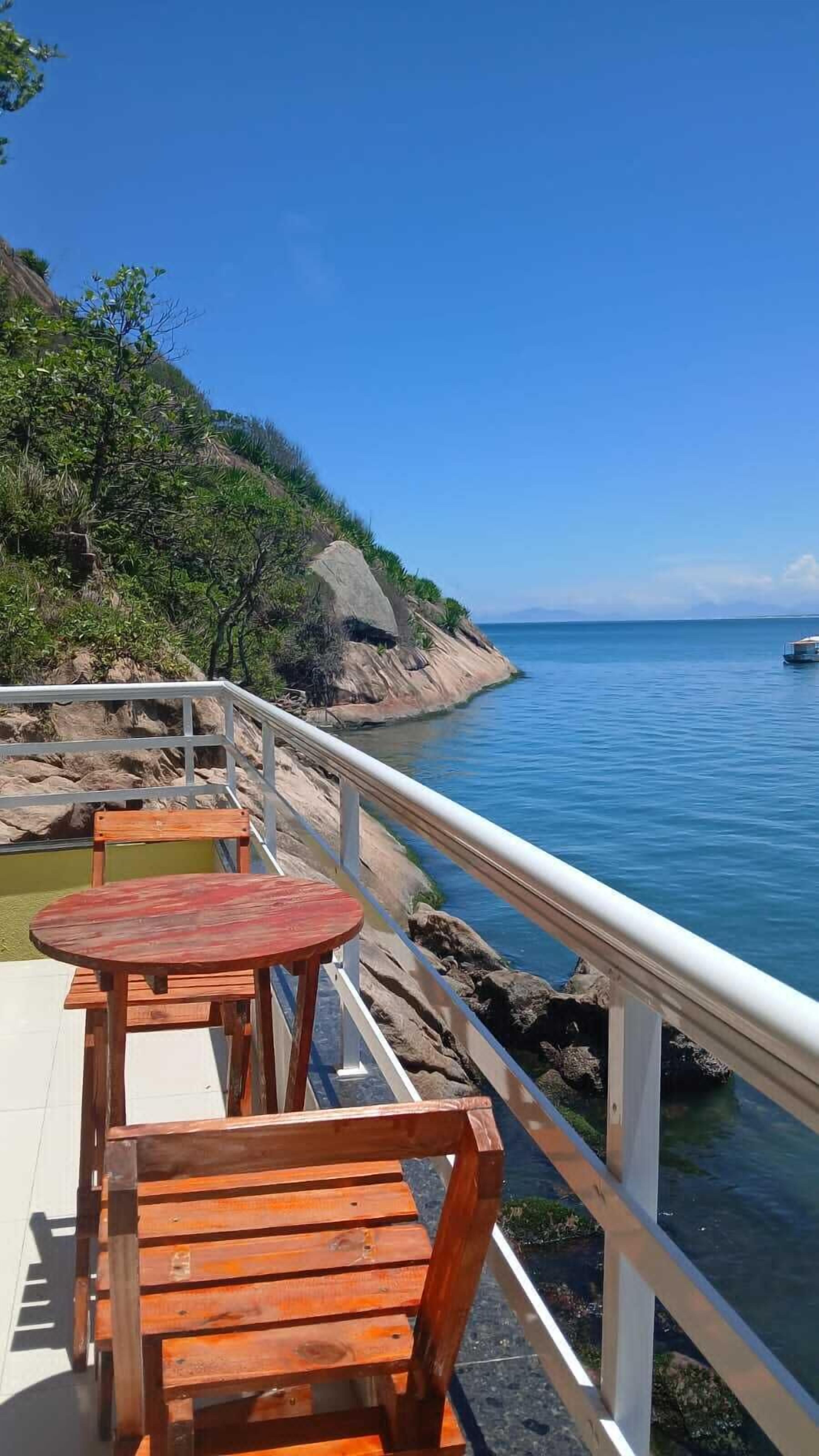 Una terraza de madera con mesas y sillas que da a un mar tranquilo, con terreno rocoso y vegetación a la izquierda.