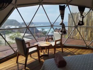 Ein Zimmer mit einem großen Fenster, das einen Blick auf den Ozean und eine Bambusumzäunung bietet. Zwei Holzstühle und ein kleiner Tisch befinden sich im Raum.