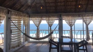 A room with a thatched roof, large windows with curtains, a hammock, and a table with chairs. - New ad (Zipolite)