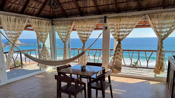 A wooden deck with a table and chairs, overlooking a beach with the ocean and a thatched roof structure. - New ad (Zipolite)