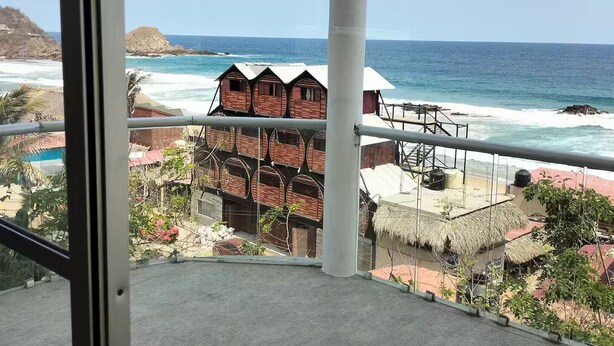 A balcony with a view of the ocean, beach huts, and a building with a thatched roof. - Grecas del mar house. (Zipolite)