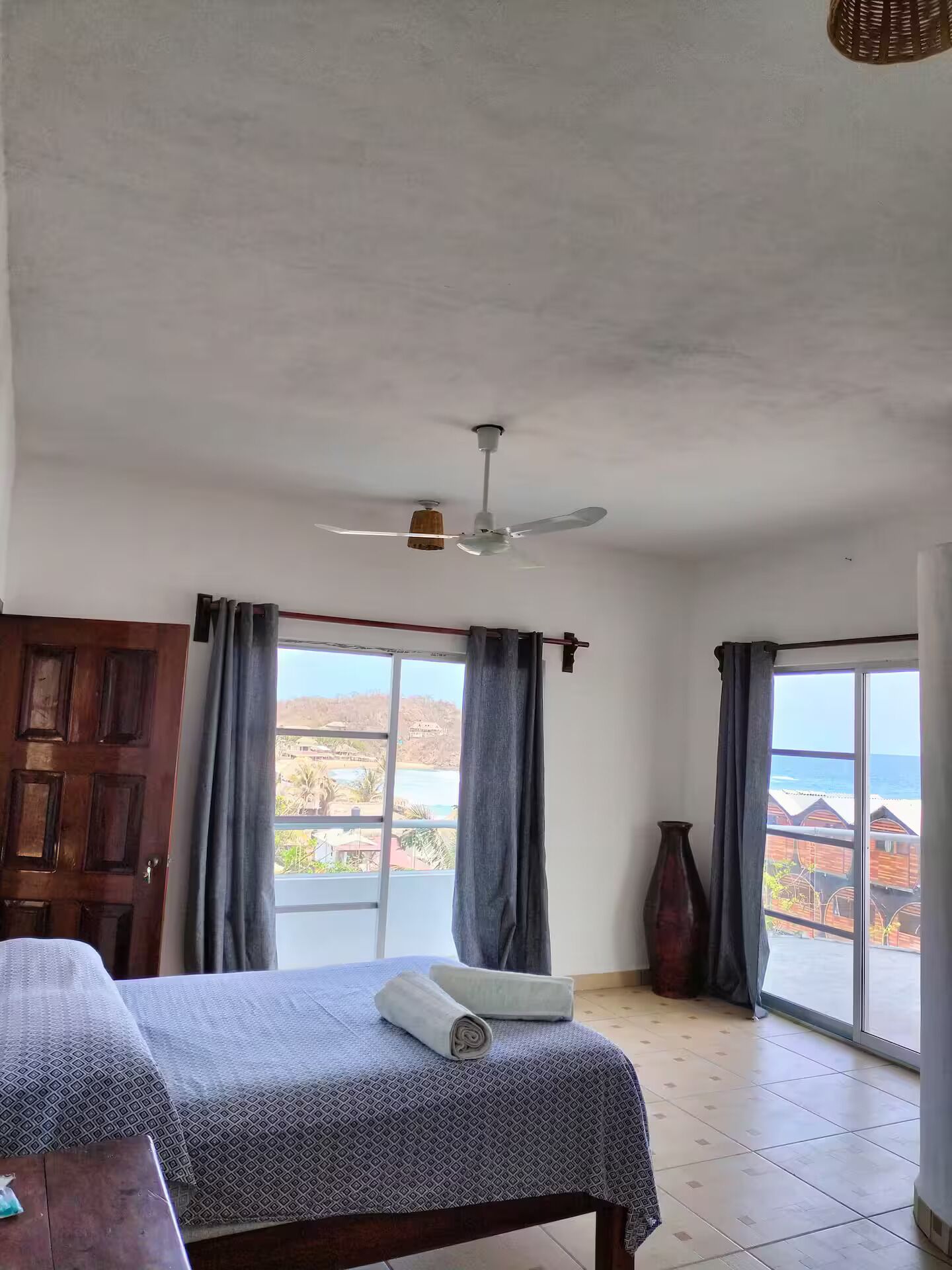 Une chambre avec un lit, un ventilateur de plafond, une vue sur la mer et une porte en bois.
