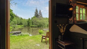 A room with a large window overlooking a pond and greenery, a wooden chair, and a television on the wall.