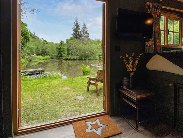 A room with a large window overlooking a pond and greenery, a wooden chair, and a television on the wall.