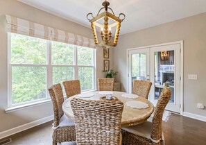 A dining area with a round table and six wicker chairs, a chandelier, and large windows with a view of greenery. - Anchor & Oar Cottage (Tullahoma)