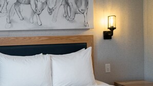 A hotel room with a bed, white pillows, a headboard, a wall-mounted light fixture, and a painting of cows.