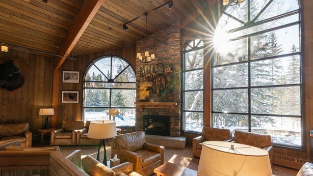 L’intérieur chaleureux d’un chalet avec des murs en bois, une cheminée en pierre, des canapés en cuir et de grandes fenêtres donnant sur des arbres couverts de neige.
