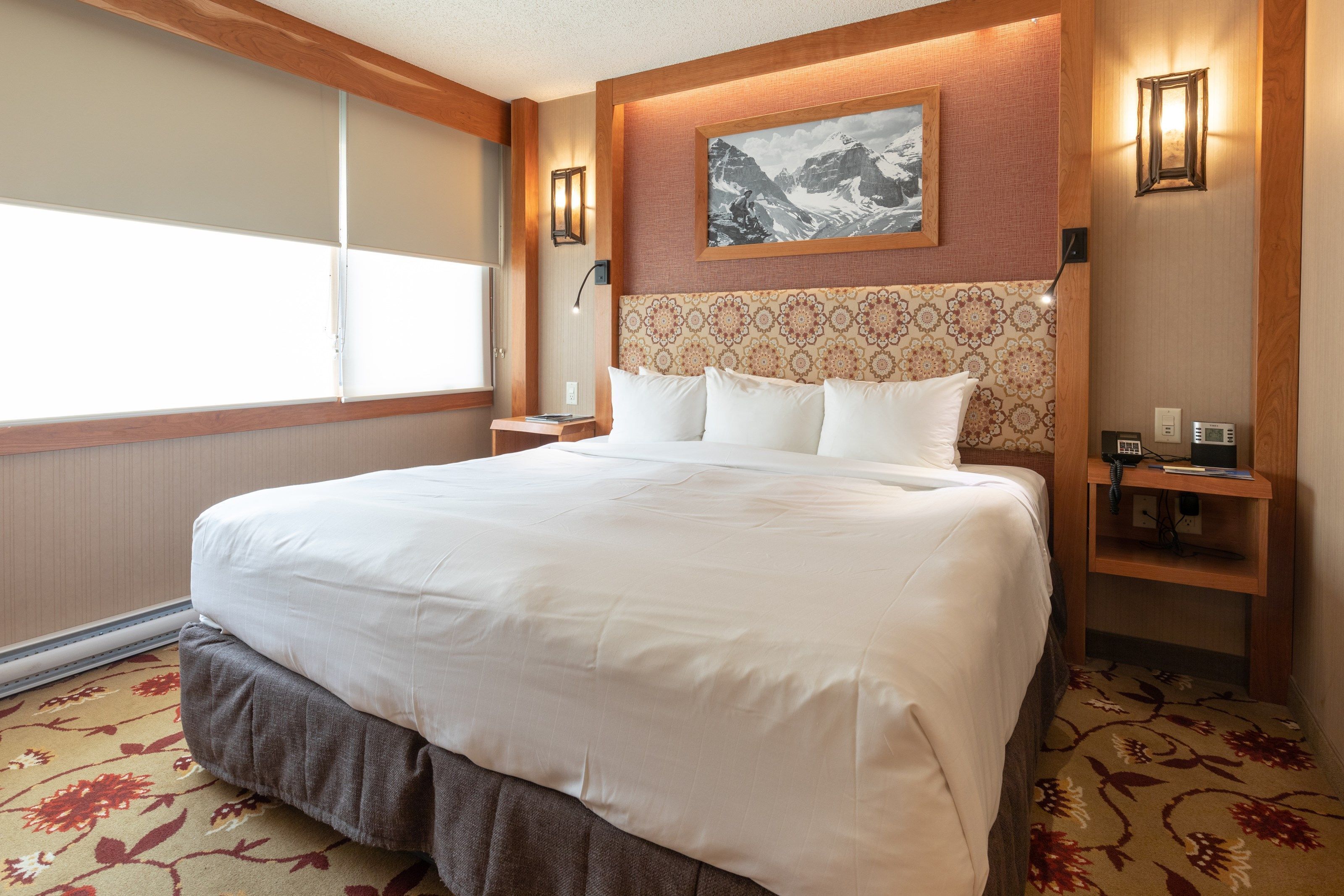 Una habitación de hotel con una cama grande, un cabecero con diseño decorativo, un cuadro sobre la cama y una mesita de noche con una lámpara.