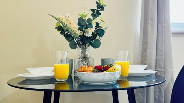 Una tavola imbandita per la colazione, con due bicchieri di succo d'arancia, una ciotola di frutta e un vaso di fiori.