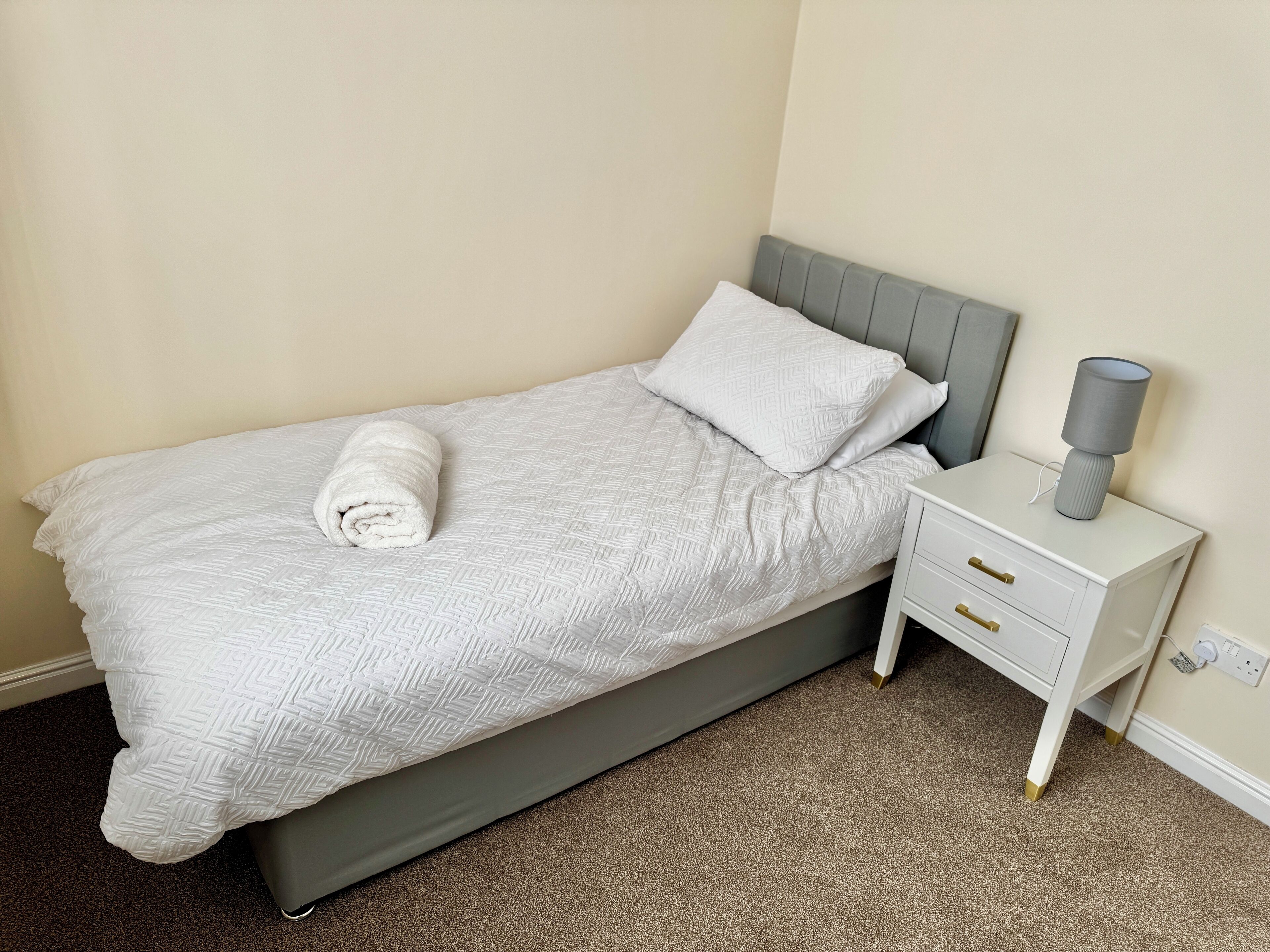 A single bed with a grey headboard, a white pillow, and a rolled white towel on the bed. There is a bedside table with a lamp and a drawer. The room has beige walls and a carpeted floor.