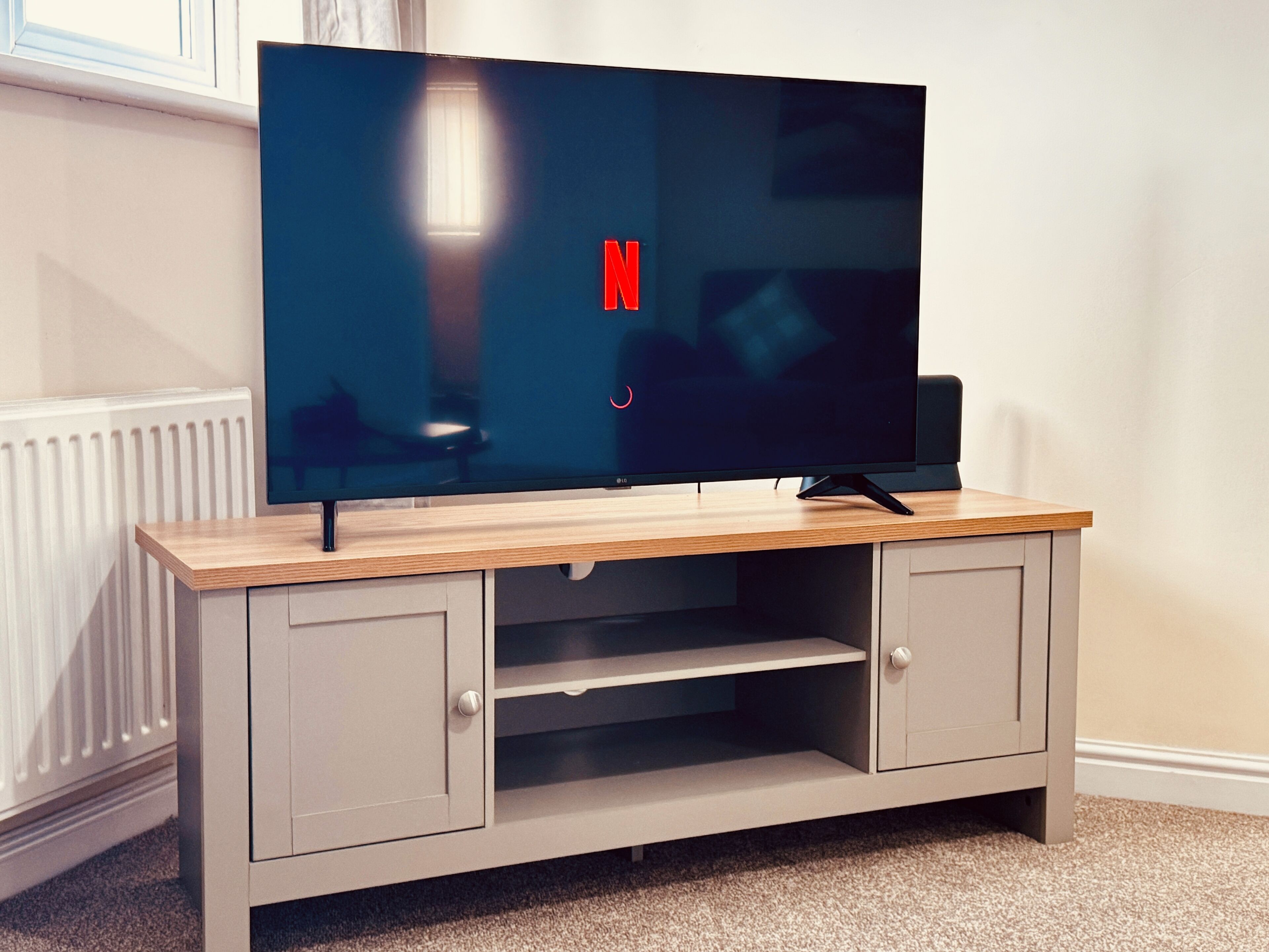 Litteänäyttöinen televisio on sijoitettu puiseen TV-telineeseen. Näkyvissä on myös Netflix-logo, patteri ja ikkuna.