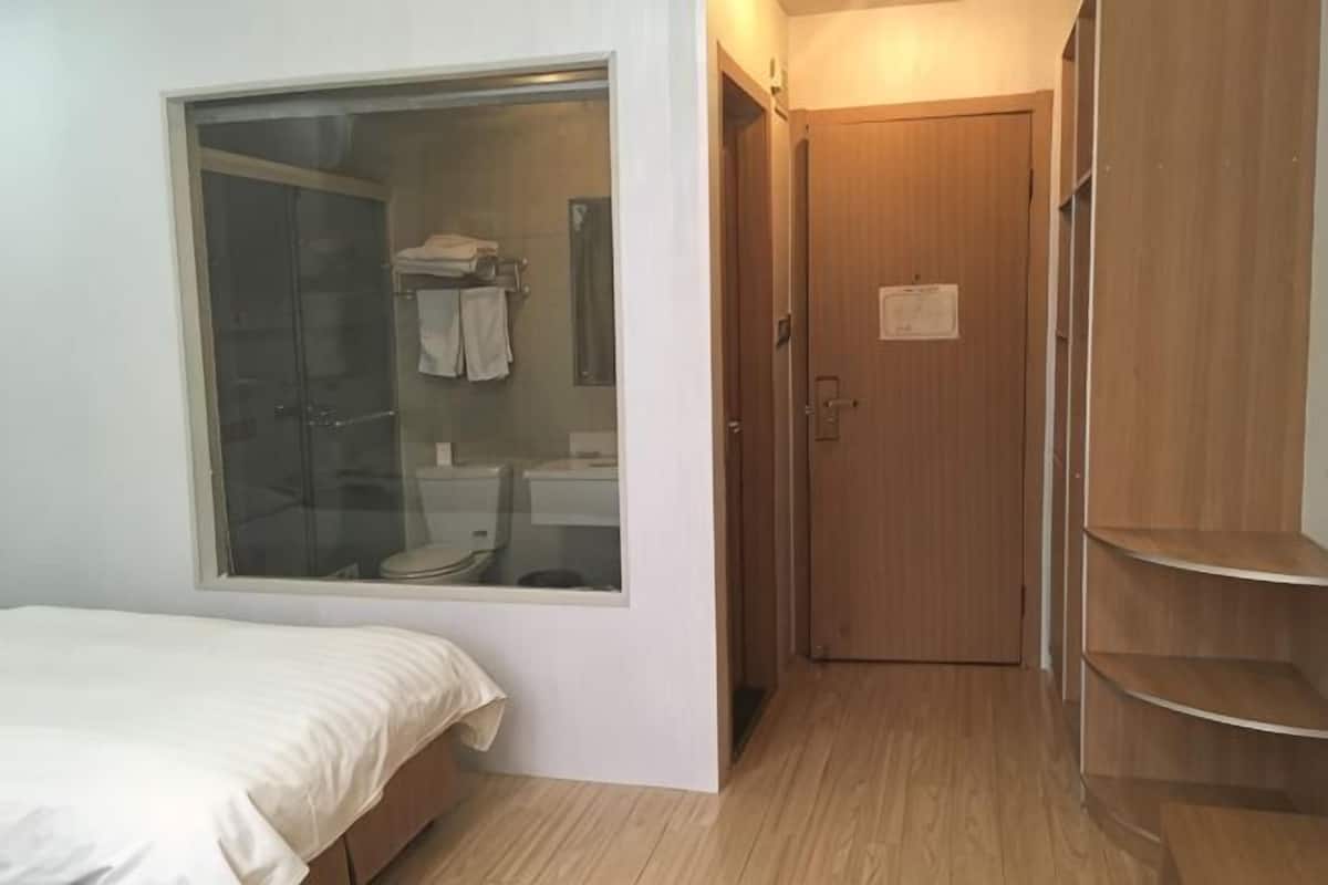 A bedroom with a large window, a wooden door, and a small bathroom visible through the window.