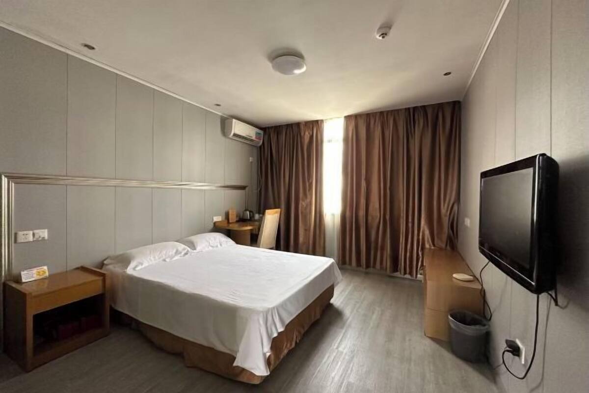 A hotel room with a large bed, a desk, a chair, a TV, and an air conditioning unit.