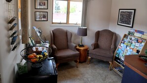 A living room with two brown armchairs, a small wooden table with a lamp, a bulletin board with travel brochures, and a window with curtains. - Marcus Daly Motel (Anaconda)