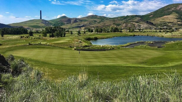 A golf course with a pond, green hills, and a tall chimney in the background. - Marcus Daly Motel (Anaconda)