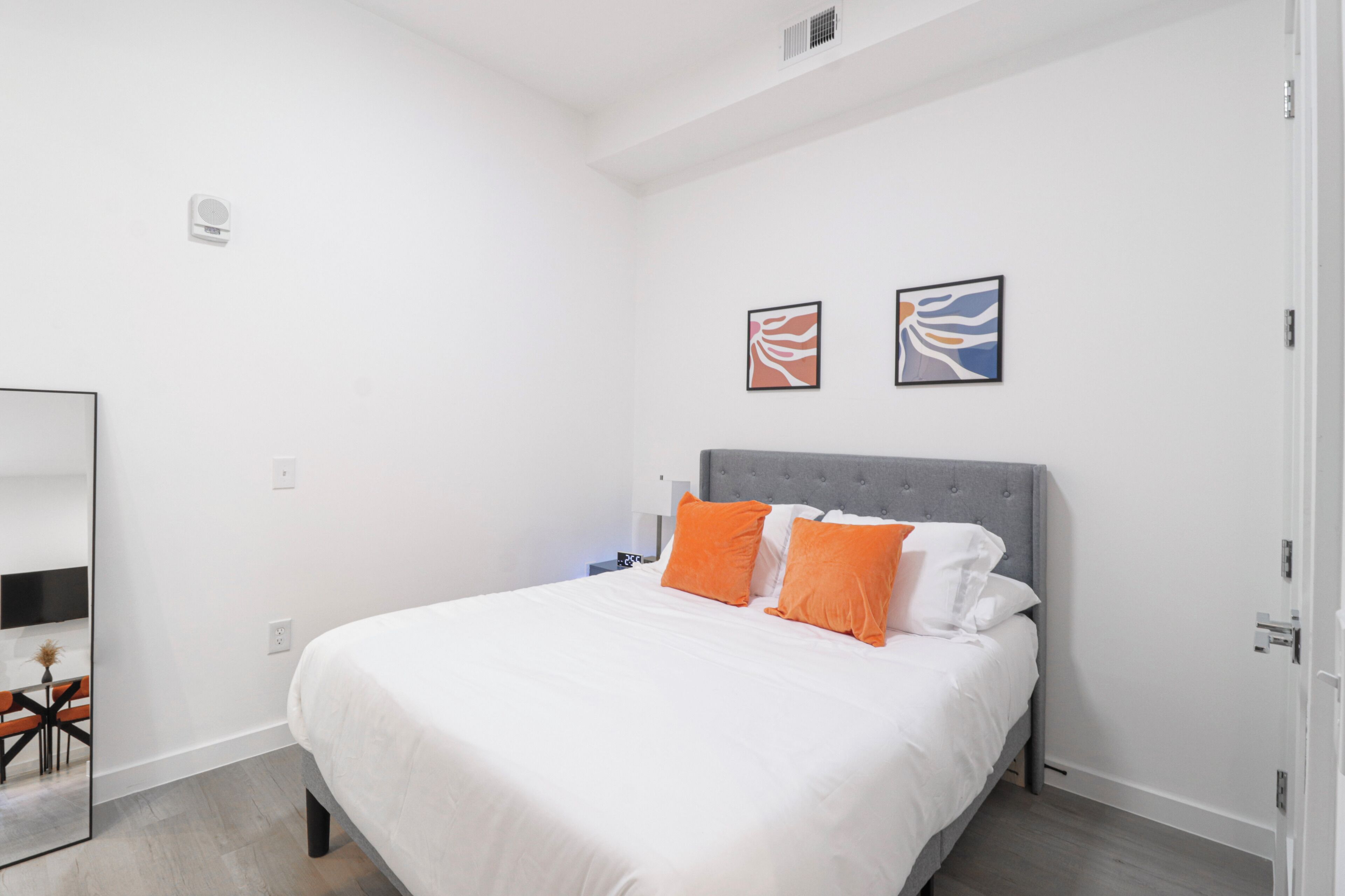 Un dormitorio con una cama grande, ropa de cama blanca y dos almohadas naranjas. Hay dos cuadros abstractos enmarcados en la pared sobre la cama.