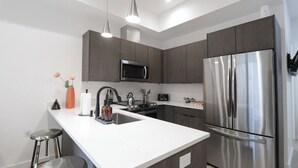 A modern kitchen with a stainless steel refrigerator, a microwave, and a sink. There are two bar stools and a vase with flowers on the counter. - Unit 4: Studio / Queen+Sofa Bed|DC Convention Centre (Washington)