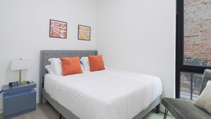 Une chambre moderne avec un lit recouvert d’un tissu gris, une literie blanche et deux coussins orange. On y trouve une table de chevet avec une lampe et une horloge numérique, ainsi que deux œuvres d’art abstraites encadrées accrochées au mur.