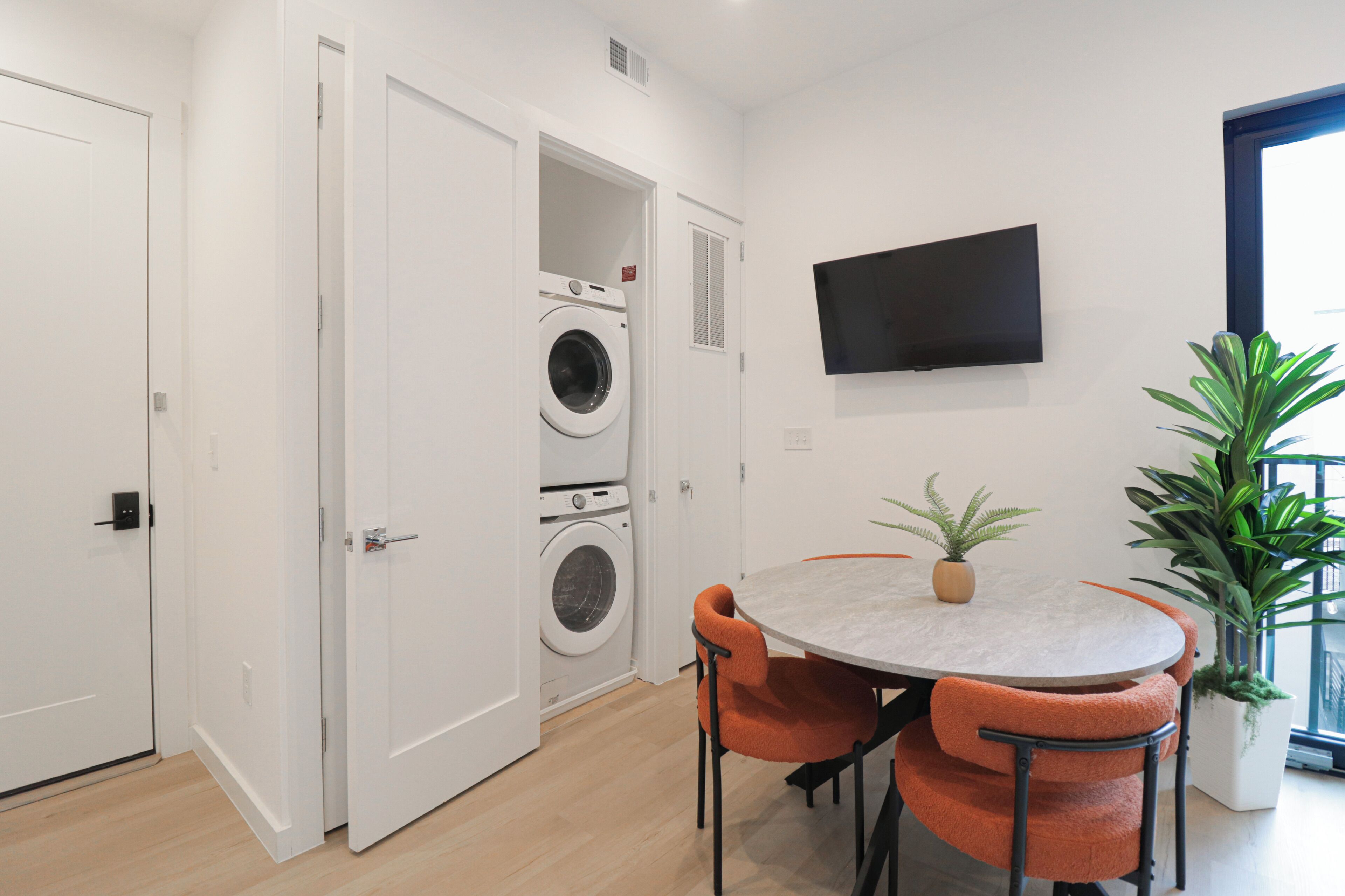 Une pièce moderne avec une table ronde, des chaises orange, un lave-linge, un téléviseur fixé au mur et une plante en pot.