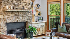 Ein gemütliches Wohnzimmer mit Kamin aus Stein, Ledersofas, Deckenventilator und großen Fenstern mit Blick ins Freie.
