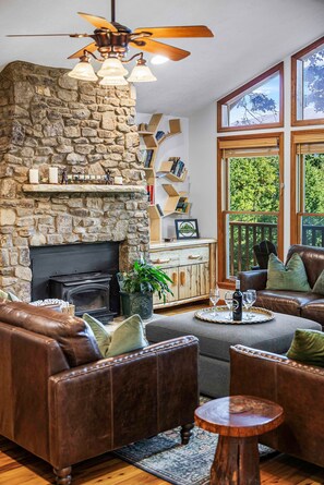 A cozy living room with a stone fireplace, leather sofas, a ceiling fan, and large windows offering a view of the outdoors. - Mountain View Woodland Respite|Hot Tub & Arcade & Pool (Sevier County)