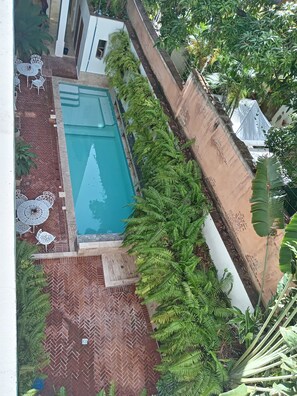 Een zwembad op het dak met een terras in een mozaïekpatroon, omgeven door weelderige begroeiing en witte tuinmeubels.