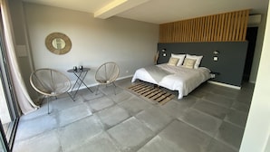 A modern bedroom with a bed, two chairs, a small table, and a mirror on the wall.