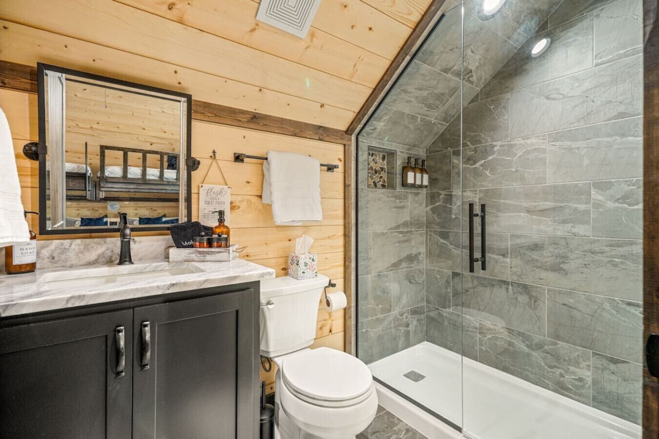 Une salle de bain moderne avec un plafond en bois, un grand miroir, un comptoir en marbre, des toilettes, une douche aux parois en verre et un petit lavabo.