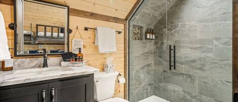 Ein modernes Badezimmer mit Holzboden, einem großen Spiegel, einer Marmorplatte, einer Toilette, einer Dusche mit Glaswänden und einem kleinen Waschbecken.