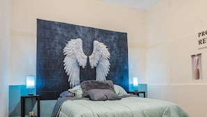 Une chambre avec une grande fresque murale représentant des ailes d’ange, une couvre-lit verte, deux lampes de chevet et un petit tableau encadré accroché au mur.