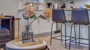 Una cocina moderna con armarios blancos, una zona de comedor con dos taburetes de bar y una mesa redonda con un jarrón y una botella de vidrio.
