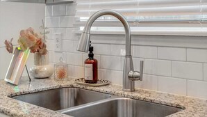 Una cocina con un lavamanos de acero inoxidable, salpicadero de azulejos blancos y una ventana con persianas.