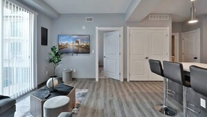 A modern living room with a large TV, a dining area with bar stools, and a kitchen with white cabinets.