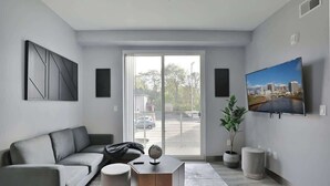 A modern living room with a grey sofa, a coffee table, a TV mounted on the wall, and a large window with a view of a parking lot.