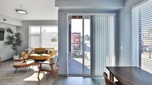 A modern living room with a sofa, coffee table, and a dining table. There is a sliding glass door leading to a balcony. - The South Campus Suite (Columbus)