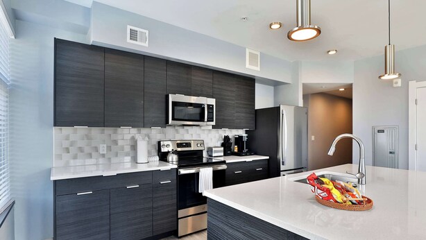 A modern kitchen with dark cabinetry, a stainless steel microwave, and a large island with a basket of snacks. - The South Campus Suite (Columbus)