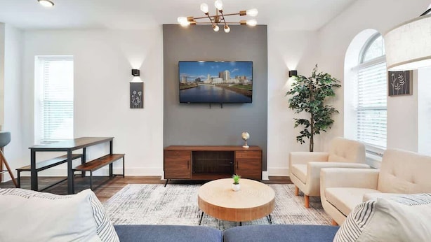 A modern living room with a flat-screen TV mounted on the wall, a round wooden coffee table, a sofa, and two armchairs. - Contemporary Vintage Home (Near Downtown!) (Columbus)