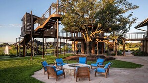 Un espace patio avec des sièges d’extérieur et un foyer, entouré d’une terrasse en bois et d’un grand arbre.