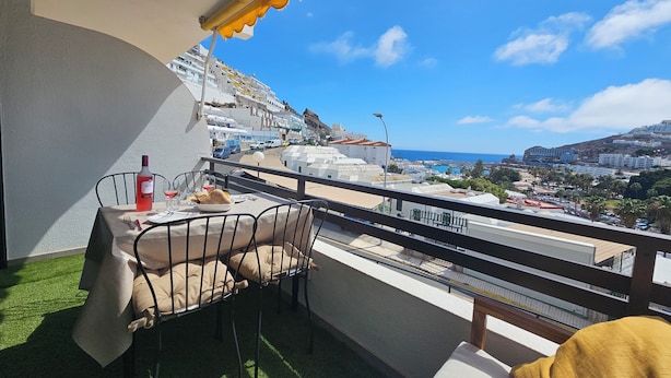A balcony with a table set for two, a bottle of wine, and a glass of red wine. The balcony overlooks a coastal town with buildings and a view of the sea. - Modern Apartment With Sea View With Pool Puerto Rico (Mogan)