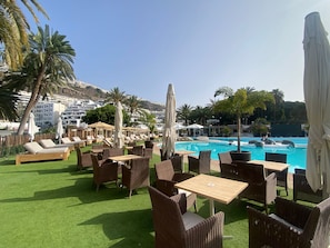 Un área junto a la piscina con sillones de descanso y mesas, piscina y palmeras.