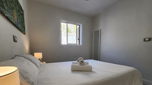 A bedroom with a large bed, a bedside table with a lamp, a window with a view of greenery, and a door. - Modern Apartment With Sea View With Pool Puerto Rico (Mogan)