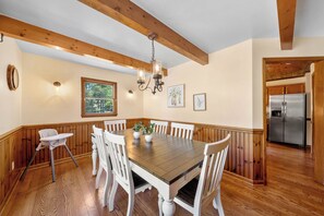 A dining area with a wooden table, white chairs, a chandelier, and a kitchen visible through an open door.