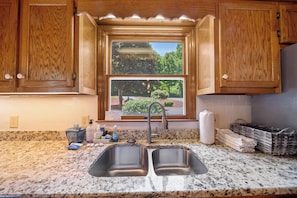 A kitchen with granite countertops, double stainless steel sinks, wooden cabinets, and a window with a view of greenery.